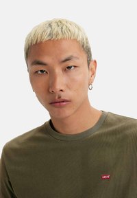 Ung person med kort, blegnet hår, iført guldøreringe og en olivengrøn Levi's skjorte med et lille rødt logo-applikation.