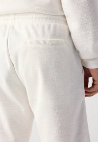 Hvite teksturerte joggebukser med et subtilt mønster, elastisk midjebånd og tilpassede lommer. Stoffet ser mykt og lett ut.