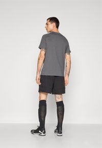 Grå t-shirt, sorte shorts, sorte knæstrømper, sorte sportssko. Synlige tatoveringer på armene, enkelt design og atletisk pasform.