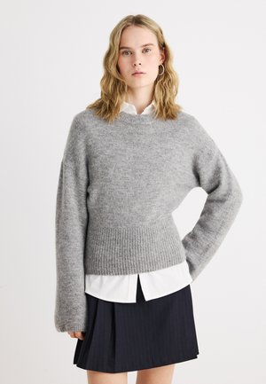 Tânără cu păr blond ondulat, purtând cercei cerc, un pulover de lână gri peste o cămașă albă și o fustă plisată neagră, stând cu o mână pe șold.