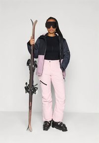 Sieviete melnā un rozā slēpošanas tērpā turēt slēpes stāvus, ģērbusies melnās slēpošanas zābakos un valkājot lielus melnus slēpošanas briļļus, stāv priekšā vienkāršai fona.