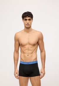 Boxers negros con cintura elástica azul que presentan la marca "TOMMY HILFIGER". El modelo está sin camiseta, mostrando un físico tonificado.