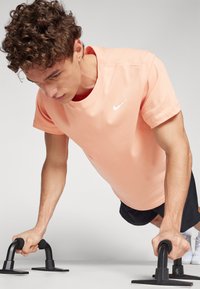 Hombre atlético realizando flexiones con mangos negros, vistiendo una camiseta de Nike de color melocotón claro y pantalones cortos oscuros; fondo sencillo.