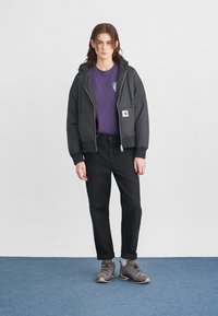 Schwarze gesteppte Jacke mit Kapuze, silbernem Reißverschluss und Logopatch, getragen über einem lila T-Shirt und schwarzen Hosen, mit grauen Wanderschuhen.