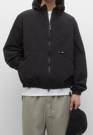 Persoon draagt een zwart gewatteerd jack met capuchon en ritssluiting, wit ondershirt, beige broek met trekkoord, en houdt een zwarte pet in de hand.