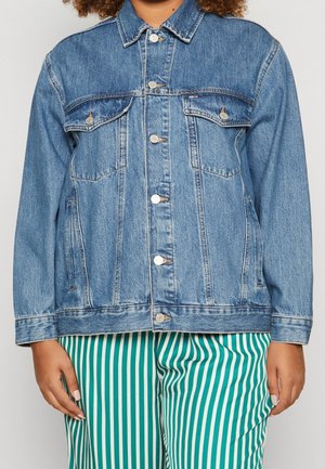 Kvinde iført en blå denimjakke med knaplukning og grønne og hvide bukser med lodrette striber, stående mod en neutral baggrund.