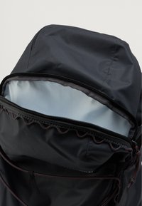 Črn najlon nahrbtnik z zadrgo, ki odkriva svetlo sivo notranjo oblogo ter rdeče šive ob zadrgi.