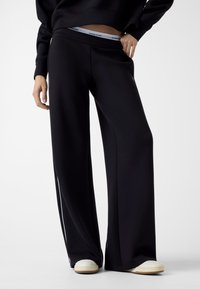 Czarne szerokie spodnie dresowe wykonane z miękkiego materiału, z regulacją w talii na sznurek i logo na elastycznej gumie. Połączone z białymi butami.