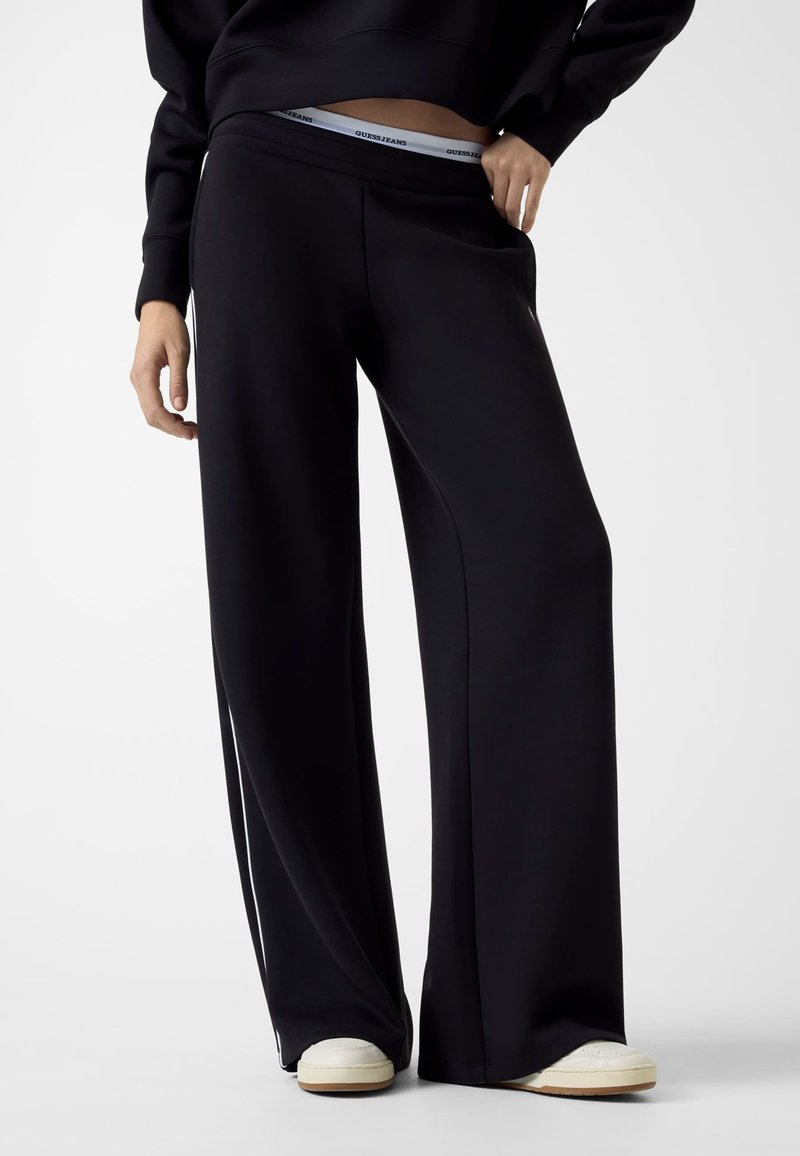 Czarne szerokie spodnie dresowe wykonane z miękkiego materiału, z regulacją w talii na sznurek i logo na elastycznej gumie. Połączone z białymi butami.