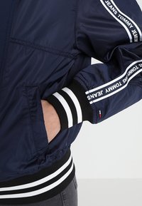 Granatowa bomberka o gładkiej teksturze, z białymi i czarnymi, prążkowanymi mankietami oraz markowymi akcentami na rękawach.