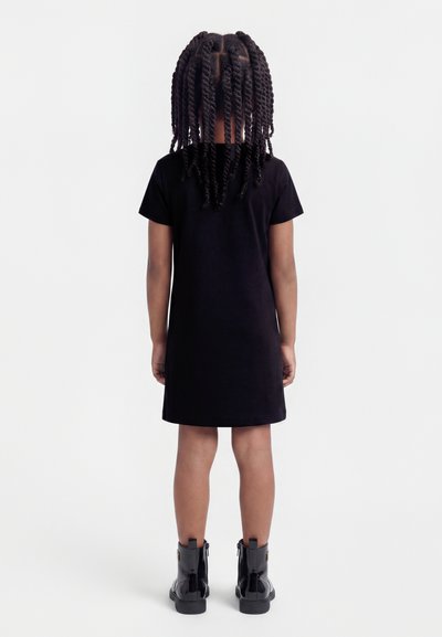 Παιδί με πλεξούδες, γυρισμένο πλάτη, φοράει μαύρο φόρεμα με κοντά μανίκια και μαύρα λαμπερά μποτάκια μέχρι τον αστράγαλο, μπροστά σε απλό λευκό φόντο.