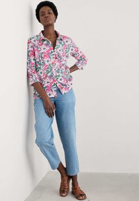 Blommig skjorta med knappar i rosa, grönt och blått på en modell, kombinerad med ljusblå denim och bruna remsandaler; avslappnad passform, trekvartsärmar.