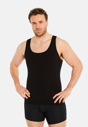 Schwarzfarbener Tank-Top aus glattem Stoff, mit Rundhalsausschnitt und weiten Armausschnitten. Das Model trägt dazu schwarze Boxershorts und hat die Hände in die Hüften gestützt.