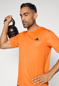 Camiseta deportiva naranja de manga corta con tejido texturizado y un logo negro en el pecho. La figura sostiene una pesa rusa negra.