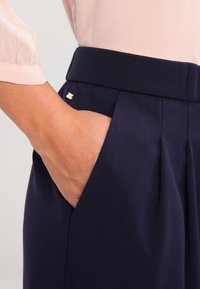 Marina-byxor med sidofickor och en texturerad midjeband; har en liten guldfärgad detalj fram, som visar en veckad design.