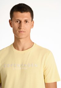 Helles gelbes Baumwoll-T-Shirt mit Rundhalsausschnitt und dem Text "COPENHAGEN LINDBERGH" in einer kontrastierenden Schriftart über der Brust.