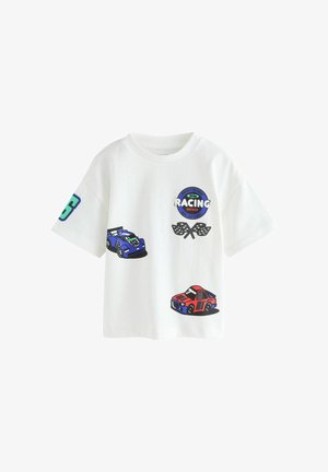 Mala beba u bijeloj majici s trkaćim automobilima, karo zastavicama i natpisom "Spreman za malog trkaćeg vozača", u kombinaciji s crnim kratkim hlačama.