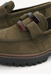 Mocassins en daim vert olive avec semelles noires épaisses, coutures détaillées et boucles rectangulaires foncées style écaille de tortue sur la bride supérieure.