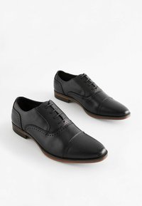 Zapatos de vestir de cuero negro con punta redondeada, acentos texturizados, detalles de brogue y un pequeño tacón apilado. Presentan un cierre con cordones.