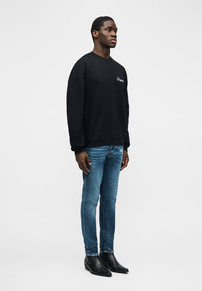 Άνδρας με μαύρο φούτερ, στενά μπλε τζιν και μαύρα μυτερά δερμάτινα μπότες, στέκεται μπροστά σε έναν απλό ανοιχτόχρωμο φόντο.