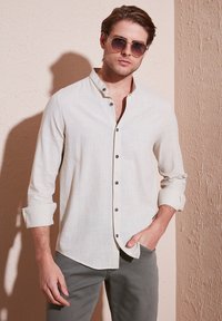 Chemise boutonnée en lin beige clair, avec un col montant, des manches retroussées et des boutons foncés, portée avec un pantalon gris. Texture lisse, design décontracté.