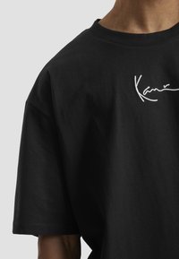 T-shirt noir à manches courtes avec un texte blanc en cursive "Kani" brodé sur la partie supérieure de la poitrine, porté par une personne devant un fond gris uni.