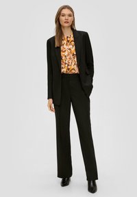 Zwart op maat gemaakte blazer en broek gedragen over een blouse met oranje en bruine patronen. Het model staat tegen een neutrale achtergrond en draagt hogere laarzen.