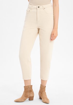 Vrouw draagt hoge, crèmekleurige cropped broeken in combinatie met tan-suède enkellaarsjes met blokhakken en ritssluitingen aan de zijkant.