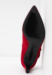 Chaussure en daim rouge à bout pointu avec semelle noire et petit talon, positionnée semelle vers le haut contre un fond blanc dans un coin.
