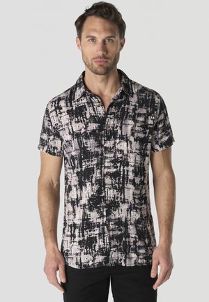 Hombre con cabello corto y oscuro, que lleva una camisa de manga corta abotonada con un patrón abstracto en blanco y negro sobre un fondo claro.