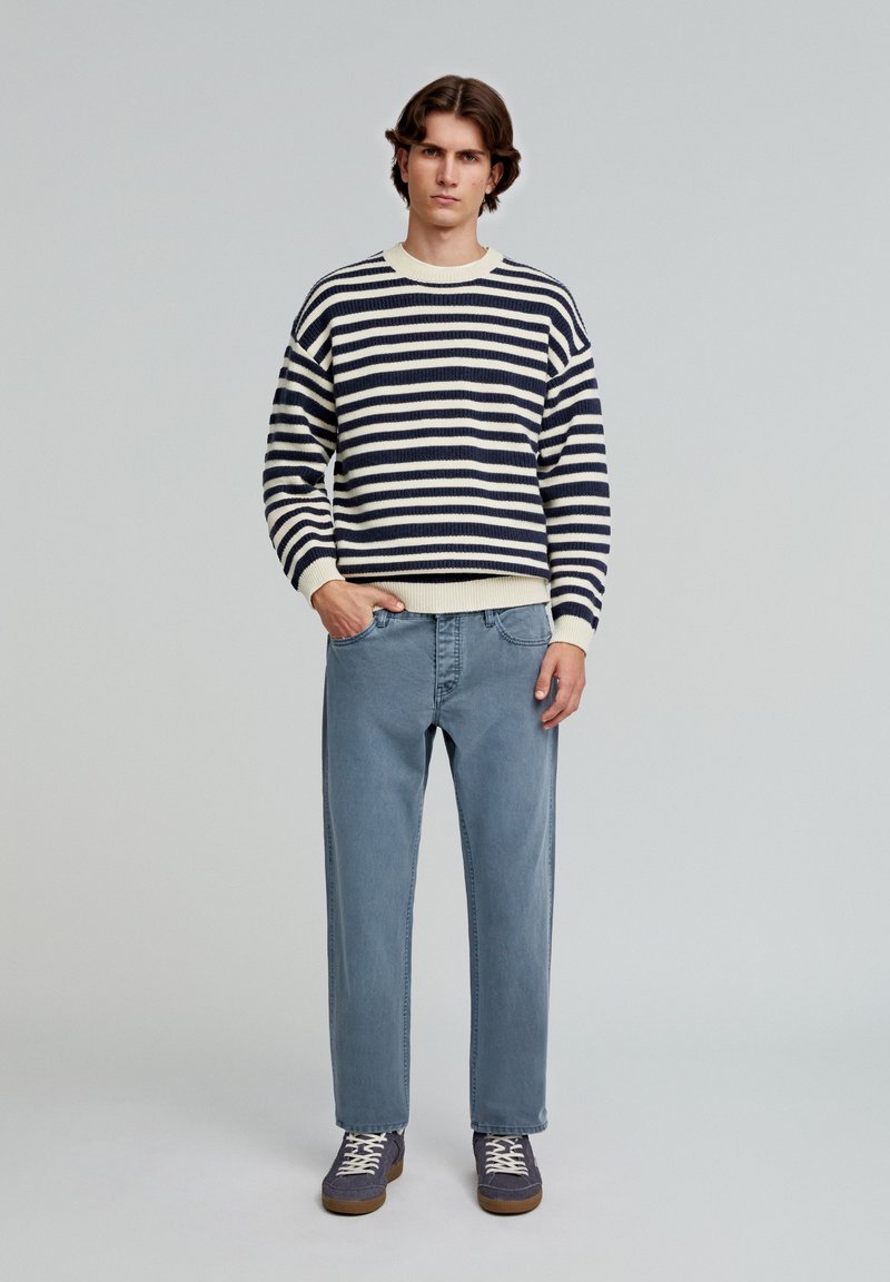 Gestreepte trui in marineblauw en crème, met een ronde halslijn, gecombineerd met lichtblauwe jeans en donkerblauwe schoenen met beige veters.