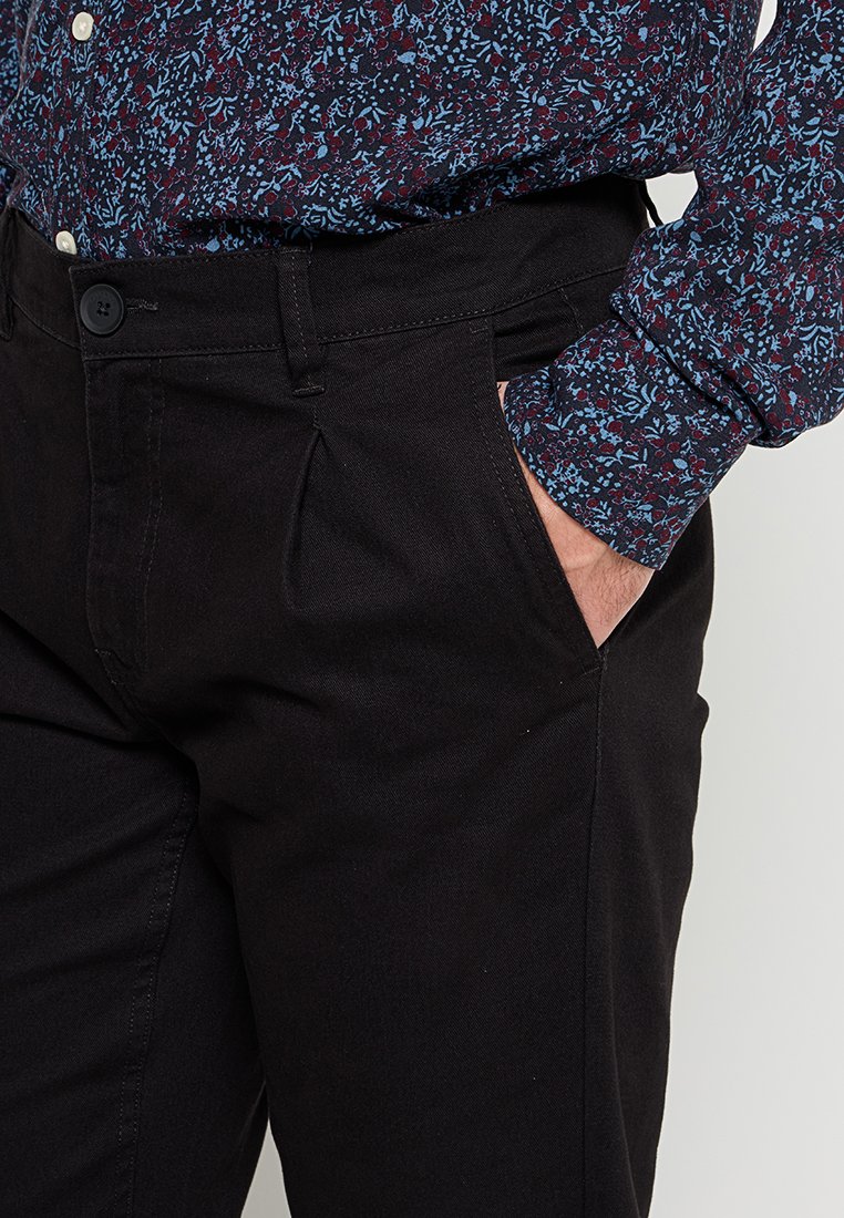 Person, der schwarze Hosen trägt, mit der Hand in der Tasche und einem Langarmshirt mit blauen und roten Blumenmustern.