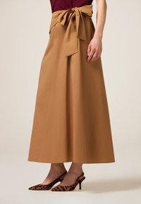 Langer, taupefarbener Rock mit einer Schleife an der Taille, kombiniert mit Slingback-High Heels im Leopardenmuster. Der Stoff wirkt glatt und strukturiert.