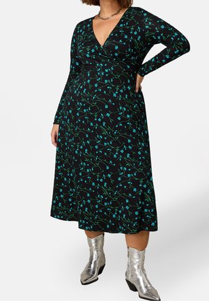 Vrouw draagt een zwarte midi-jurk met blauw bloemenpatroon, lange mouwen, V-hals, gecombineerd met zilveren laarzen tot halverwege de kuit, staat met de hand op de heup.