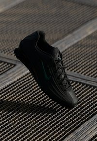 Musta urheilujalkine, jossa on turkoosi swoosh-logot, roikkuu metalliverkkoisen lattian yllä hämärässä valaistuksessa.