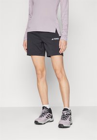 Calças curtas atléticas pretas com um logótipo na lateral, combinadas com uma blusa de manga longa lilás clara e sapatilhas de trilha cinzas e roxas.