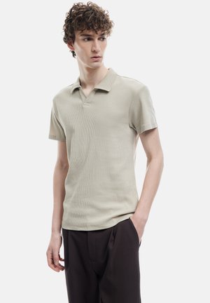 Camisa polo de color beige claro con un acabado texturizado, mangas cortas y un cuello clásico, combinada con pantalones oscuros. El modelo está de pie con las manos en los bolsillos.