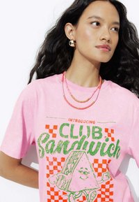 T-shirt en coton rose avec un imprimé graphique d'un personnage de sandwich, avec des lettres bold en vert et rouge et des accents à carreaux.