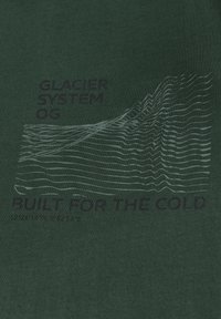 Donkergroene t-shirt met een wit golfpatroon en tekst "GLACIER SYSTEM OG" en "BUILT FOR THE COLD." Zachte katoenstructuur.