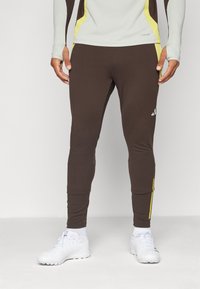 Pantalons de sport marron en tissu extensible, présentant une coupe ajustée, des accents jaunes sur les côtés et un logo sur la jambe gauche.