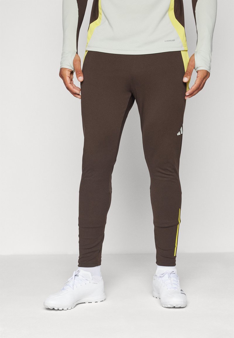 Pantalons de sport marron en tissu extensible, présentant une coupe ajustée, des accents jaunes sur les côtés et un logo sur la jambe gauche.