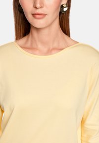 Light yellow long-sleeve top with a wide neckline, made of soft fabric. Model wears round, shiny earrings for added detail.