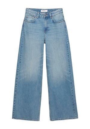 Kvinde med langt krøllet blondt hår iført en hvid tanktop og løse blå jeans, med den ene hånd i lommen og den anden, der løfter hendes skjorte.