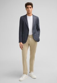 Leichter marineblauer Blazer aus strukturiertem Stoff, kombiniert mit einem weißen T-Shirt, beigen Hosen und weißen Sneakern, vor einem neutralen Hintergrund stehend.