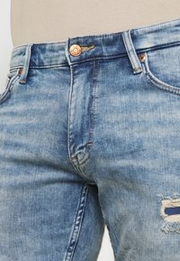 Světlé modré džínové kalhoty s opotřebovanou oblastí na stehně, měděným knoflíkem a oranžovým šitím. Vybledlá textura s jemnými variacemi.