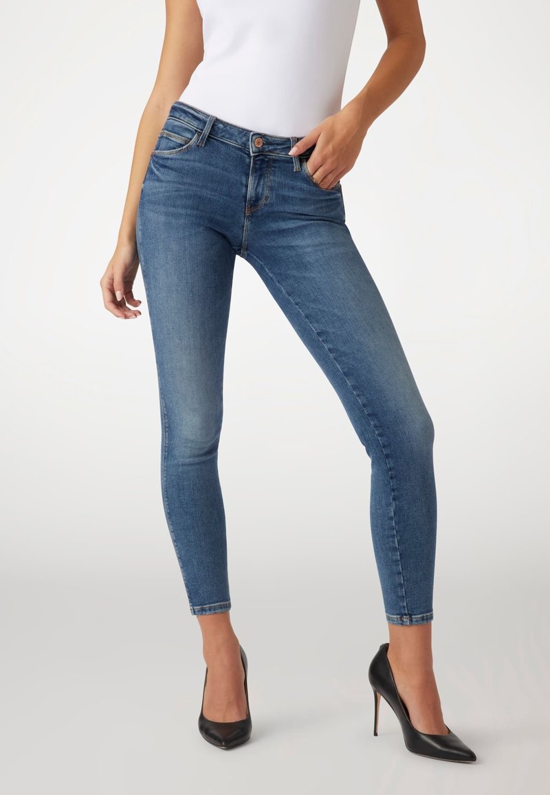 Femme portant un jean bleu moulant, un haut blanc et des chaussures à talons noires, posant avec une main dans la poche sur un fond uni.