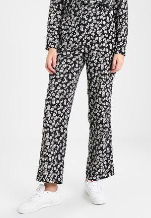 Kvinde iført sorte bukser med hvidt blomsterprint og hvide sneakers, stående foran en ensfarvet lys baggrund.