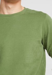 Zelená pánská košile s dlouhým rukávem vyrobená z měkké, hladké látky; má kulatý výstřih a přiléhavý střih s jemnou texturou.