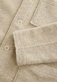 Beige vlněný cardigan s knoflíkovou přední částí, který má kapsu a ohrnutý manžet. Tkanina má jemnou texturu a subtilní jemné tečky.