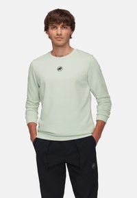 Light green sweatshirt with long sleeves, round neckline, and a black logo on the chest. Soft texture and relaxed fit. Black pants.